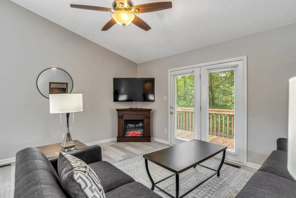 A spacious living room filled with natural light, featuring a comfortable sofa, sleek coffee table, a stylish fireplace, and a large television. Double doors offer a view to the outside, reinforcing the open-concept feel. The space is thoughtfully arranged for conversation, relaxation, and group activities.