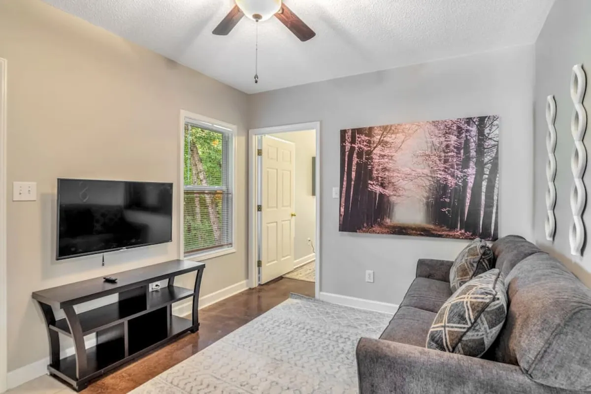 A cozy and inviting living area with a plush sofa adorned with decorative pillows, a large piece of wall art, and a television unit. Natural light streams in from a nearby window, while a doorway connects to another part of the home, emphasizing the interconnected nature of the space.