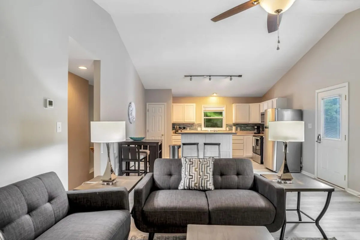 The image showcases a bright, open-concept living space with a comfortable sofa, a stylish kitchen island perfect for gathering, large windows that let in natural light, and cozy table lamps. The layout invites guests to socialize, relax, and enjoy shared meals or game nights in a contemporary setting.