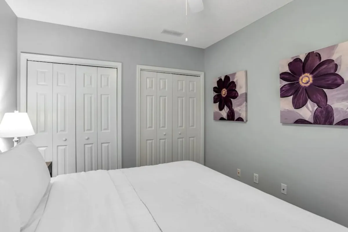 A cozy king bed draped in crisp white linens sits next to a softly-glowing bedside lamp and floral artwork, with natural light filtering in to create a serene atmosphere. The setting invites restful mornings and intimate, peaceful evenings.