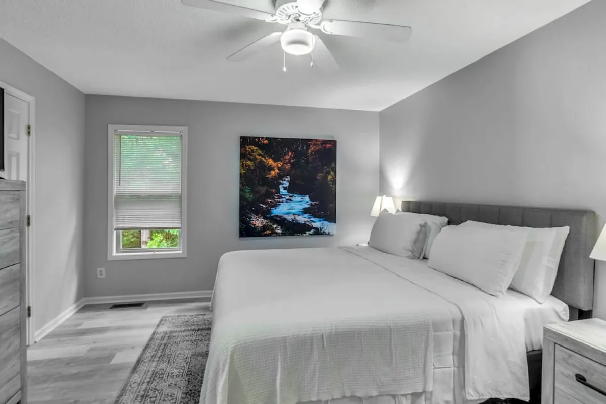 A serene bedroom scene showcasing a bed dressed in white linens, a striking large artwork depicting a natural stream above the bed, abundant natural light streaming through a window, and a modern ceiling fan above. The overall effect is peaceful and connects the interior to the outdoors through the artwork.