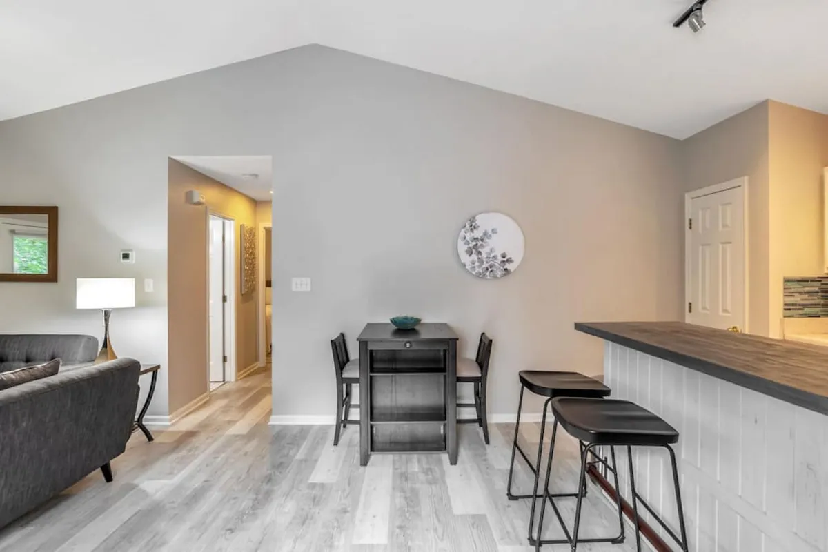 A modern, open-plan living and dining area with a stylish sofa, dining table, bar stools, and contemporary wall art. The space is arranged for socializing, casual dining, and relaxation, creating a cohesive zone for multiple group activities.