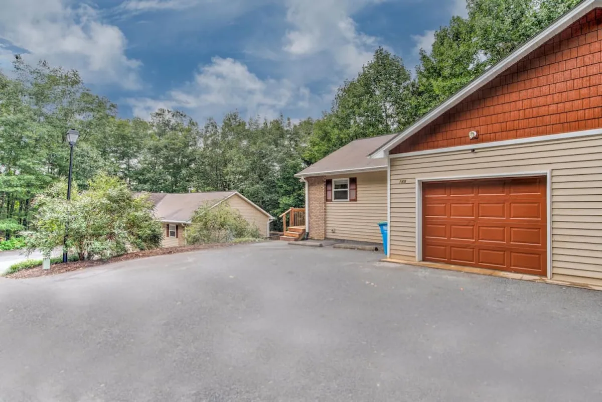 A wide-angle view of the main house exterior featuring a driveway, garage door, reserved parking area, and surrounding greenery. The scene is inviting, clearly showing both the garage and parking spaces, with easy access for guests and their vehicles.