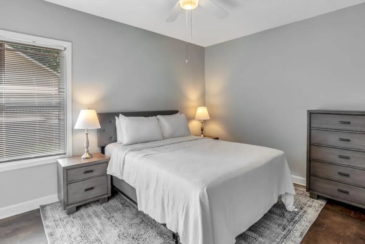 A tranquil bedroom scene featuring a plush bed, nightstand with lamp, dresser, and a window letting in gentle sunlight, evoking a personal retreat ideal for quiet moments, reading, or simply enjoying each other's company.